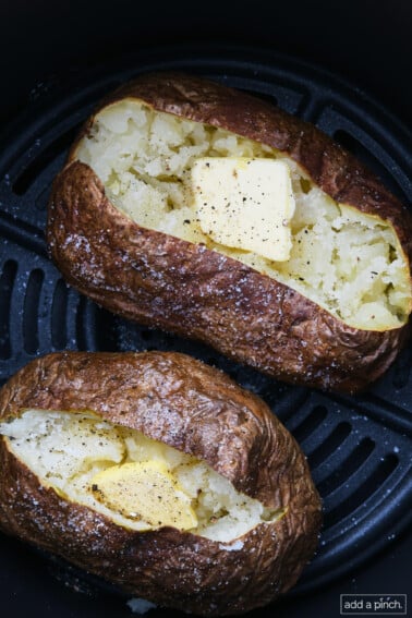 Two air fryer baked potatoes with butter, salt, and pepper in an air fryer basket.