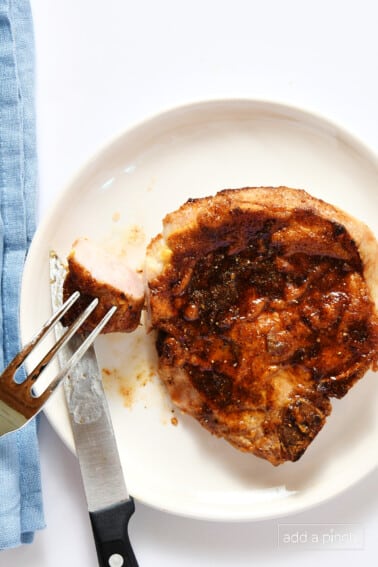 Bite of pork chop on the end of a fork with the rest of the pork chop on a white plate with a blue napkin.