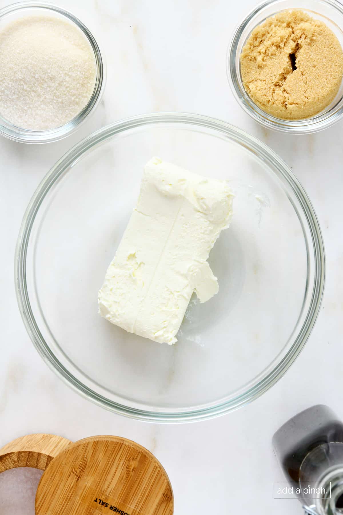 Glass bowl with block of cream cheese, surrounded by smaller glass bowls of brown sugar, sugar, salt and a glass bottle of homemade vanilla extract.