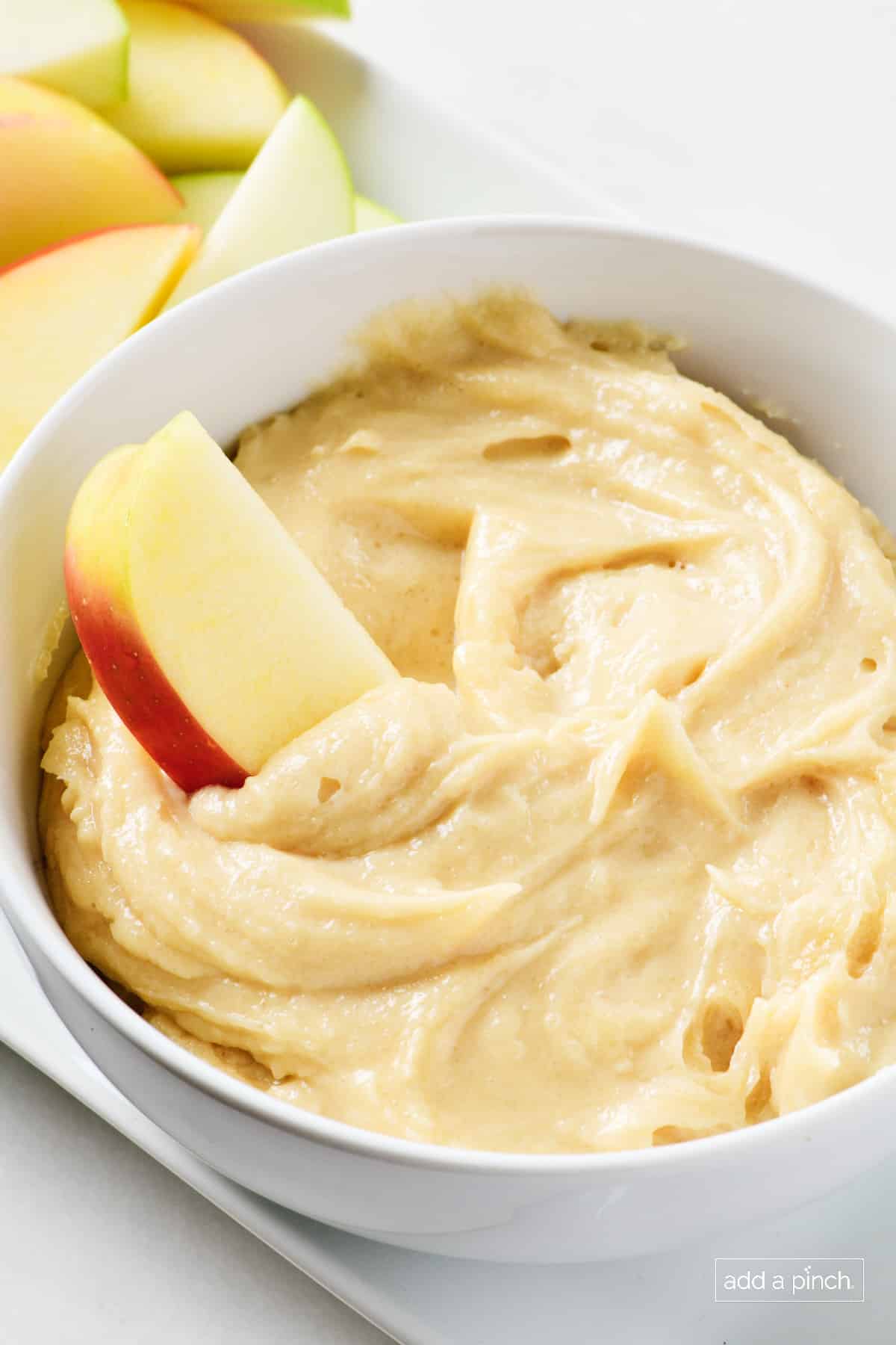 Apple slice dipped into apple dip in white bowl, surrounded with more slices. 