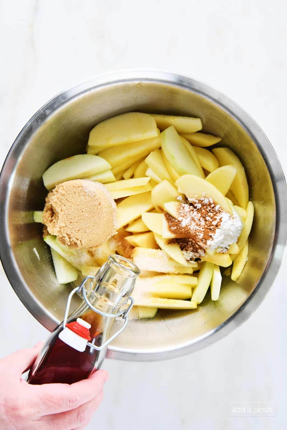 Stainless mixing bowl holds sliced apples, sugars, spices and has vanilla being added, to make apple pie.