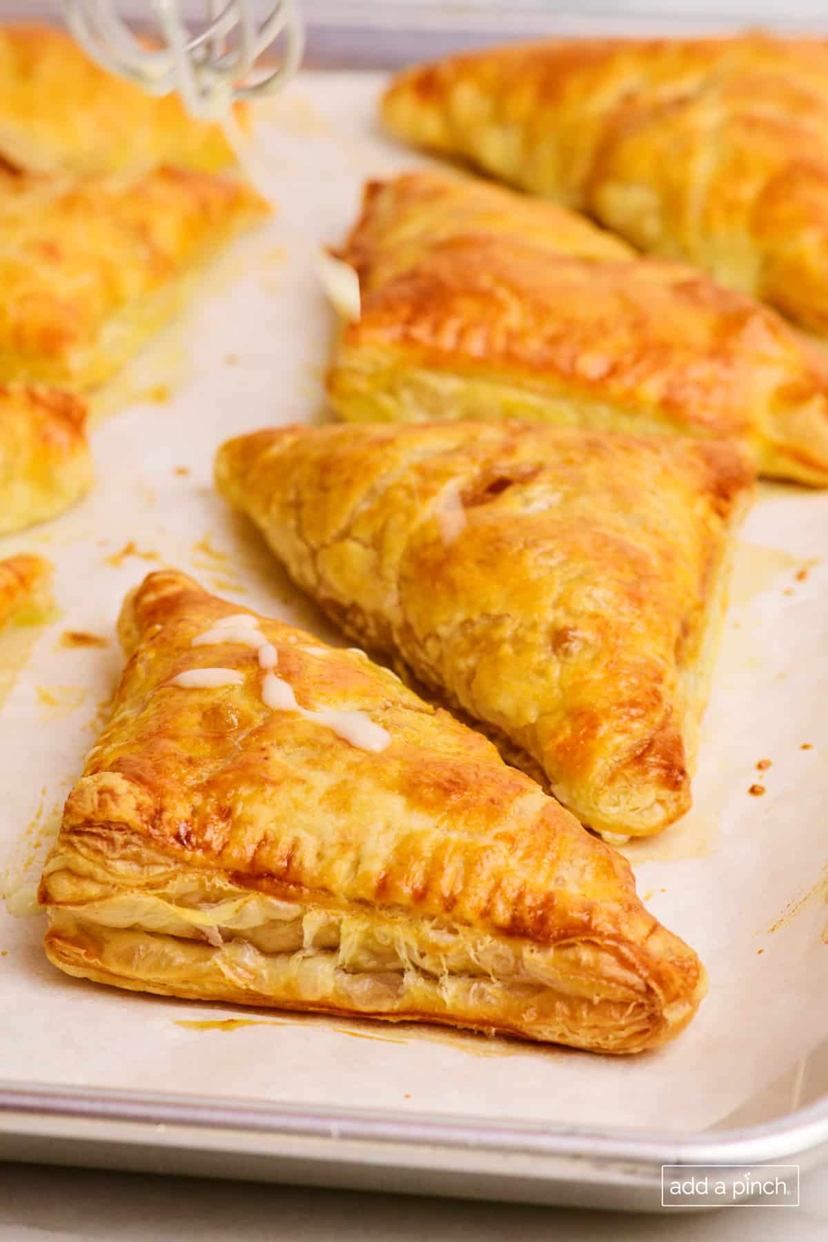 Golden brown apple turnovers on a baking sheet being drizzled with glaze.