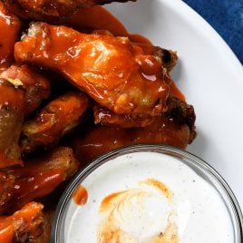 White plate holds several buffalo chicken wings coated in buffalo sauce, sitting next to a bowl of ranch dressing for dipping.