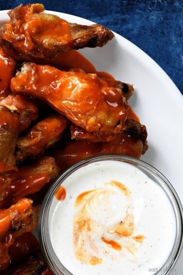 White plate holds several buffalo chicken wings coated in buffalo sauce, sitting next to a bowl of ranch dressing for dipping.