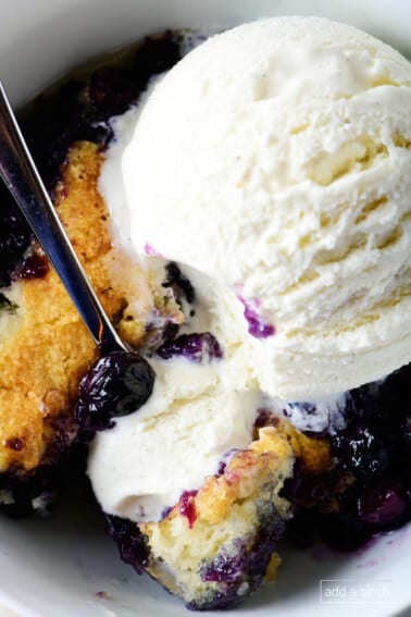 Photo of blueberry cobbler topped with vanilla ice cream in white bowl.