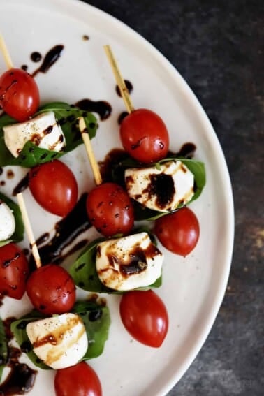 Caprese Salad Skewers of tomatoes, mozzarella, basil, drizzled with balsamic glaze on a white platter.