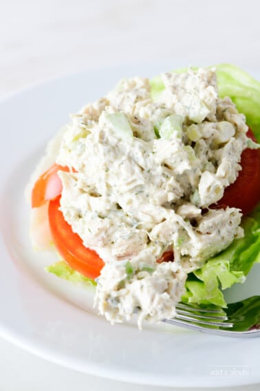 Classic chicken salad on a white plate with slice of tomato, lettuce, and a fork with a bite of chicken salad.