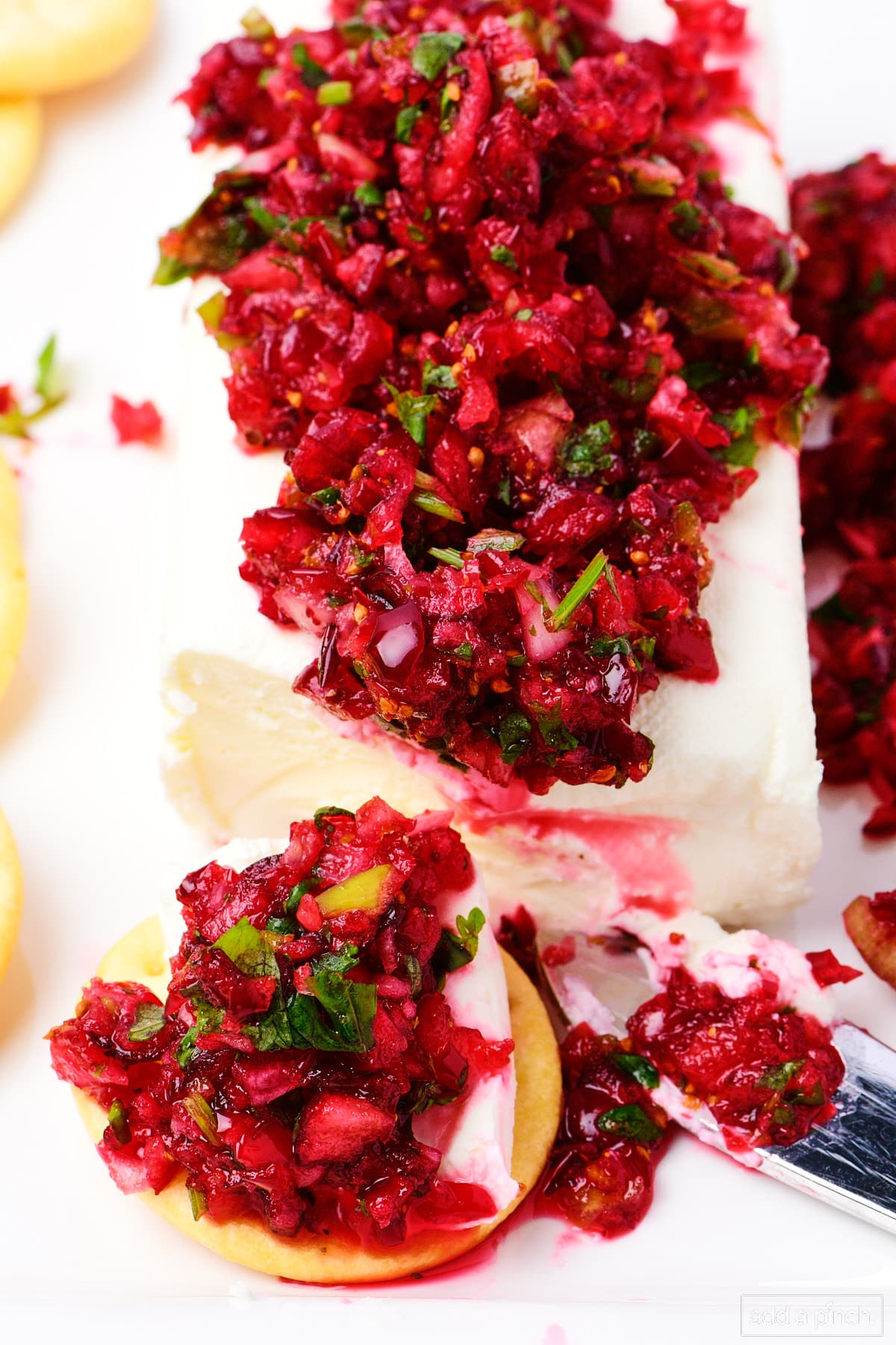 Spreader scoops off softened cream cheese topped with cranberry salsa onto a cracker. The remainder of the cranberry salsa over cream cheese is served on the white platter as well.