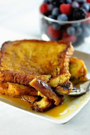 Photograph of French toast on a white plate with maple syrup and a bowl of fruit in the background.