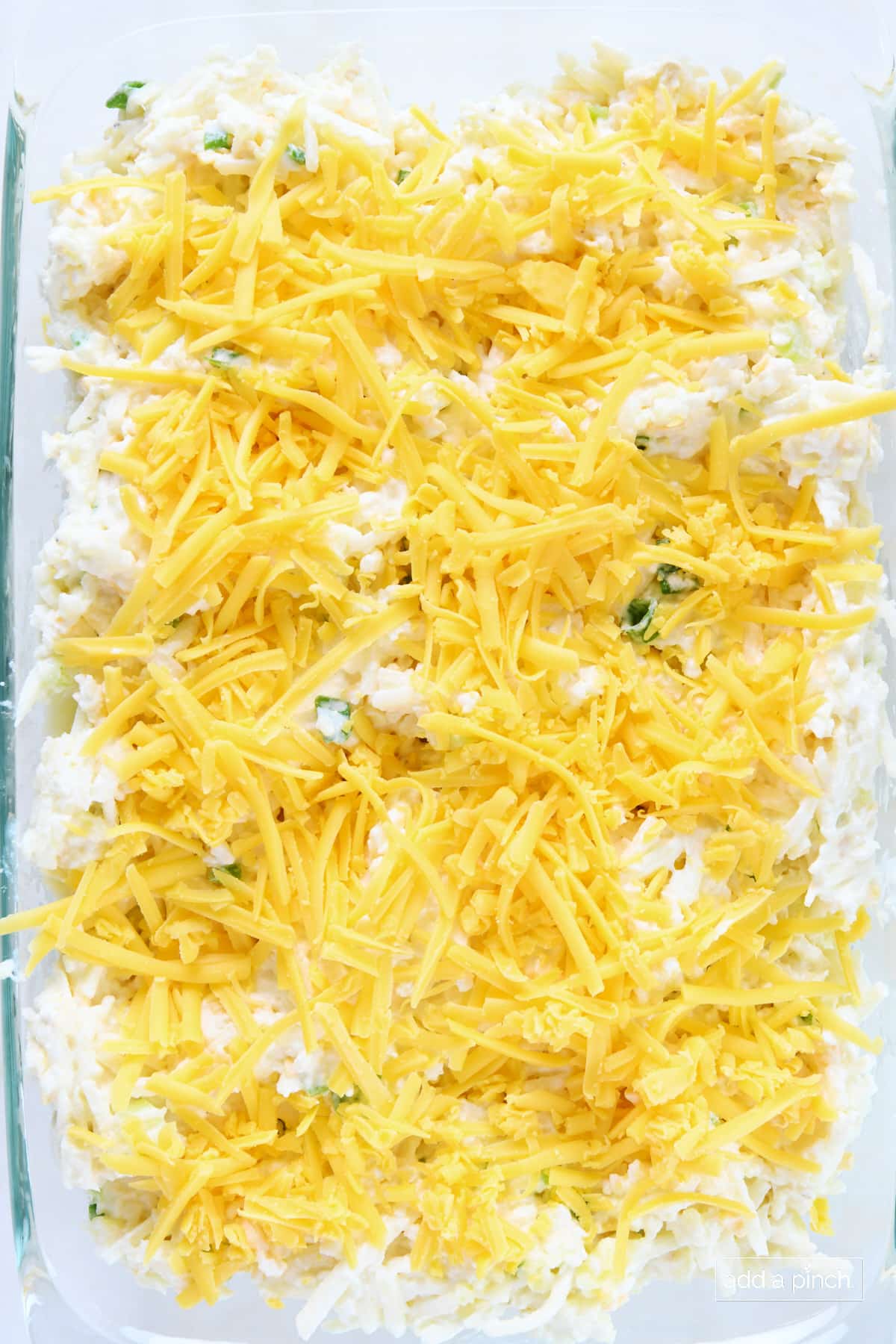 Glass baking dish of uncooked hashbrown casserole covered in shredded cheese. 