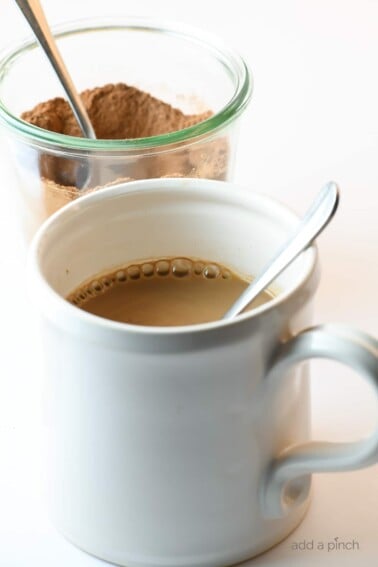 Photograph of white mug filled with hot cocoa and a clear jar of hot cocoa mix behind it on a white counter. // addapinch.com