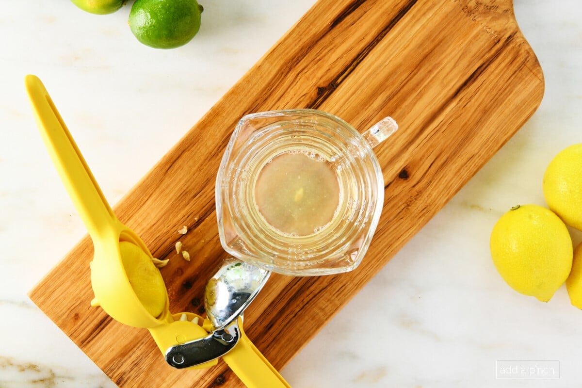 photo of yellow citrus juicer with lemon half inside and clear glass measuring cup on a cherry board sitting on a marble surface with a green lime at top left side 