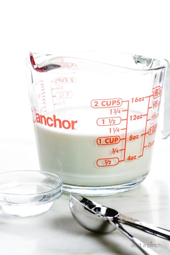 Glass measuring cup with milk, glass bowl with vinegar, and measuring spoons sit on a marble countertop.