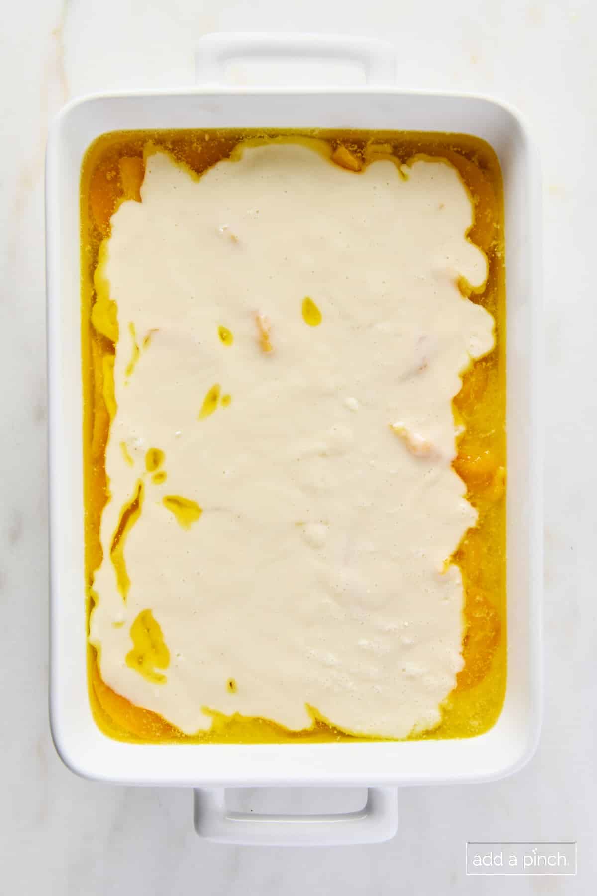 Peaches, butter and cobbler batter in a white baking dish ready to bake.