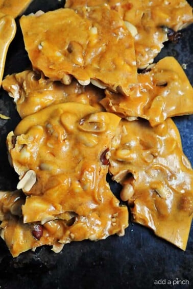 Nut filled peanut brittle pieces stacked on a dark surface.