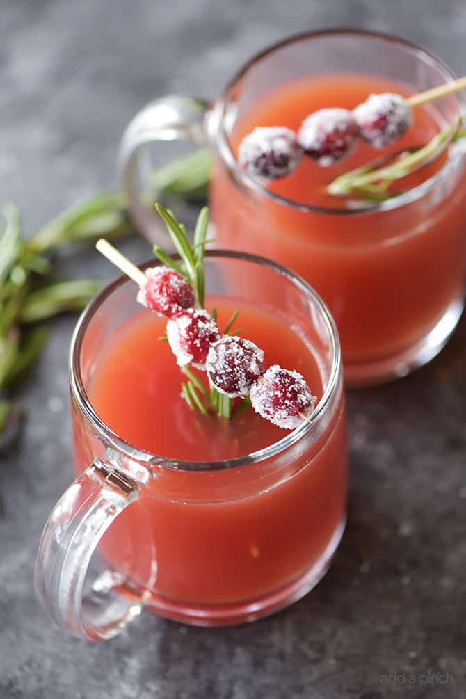 Glass mugs of warm pomegranate apple cider with garnish of rosemary sprig and skewer of sugared cranberries, all on black granite counter scattered with rosemary sprigs.