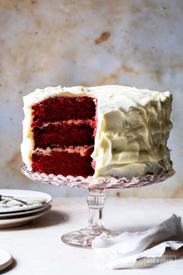 Photograph of red velvet cake with white frosting with a slice cut so that you can see the three layers of the rich, velvety cake.