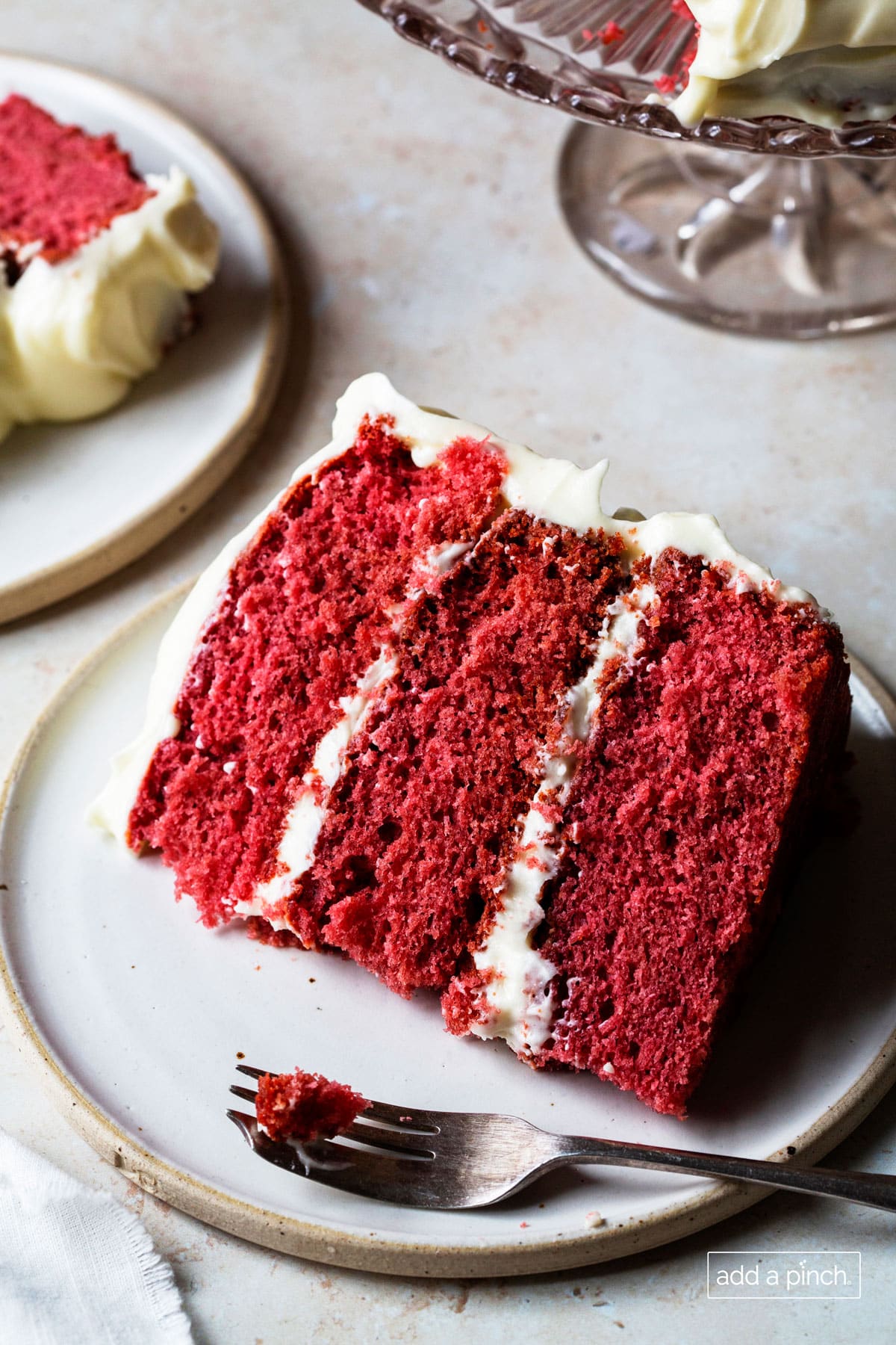 Slice of red velvet cake sitting close to cake plate and another slice nearby.