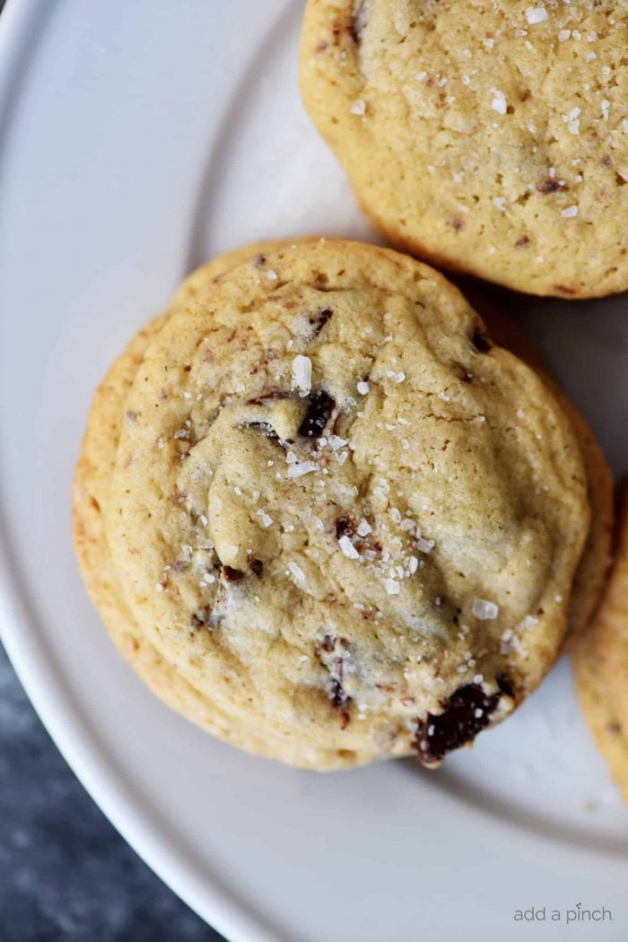 Photograph of chocolate chip cookie topped with flakes of salt on a white plate on a dark background. 