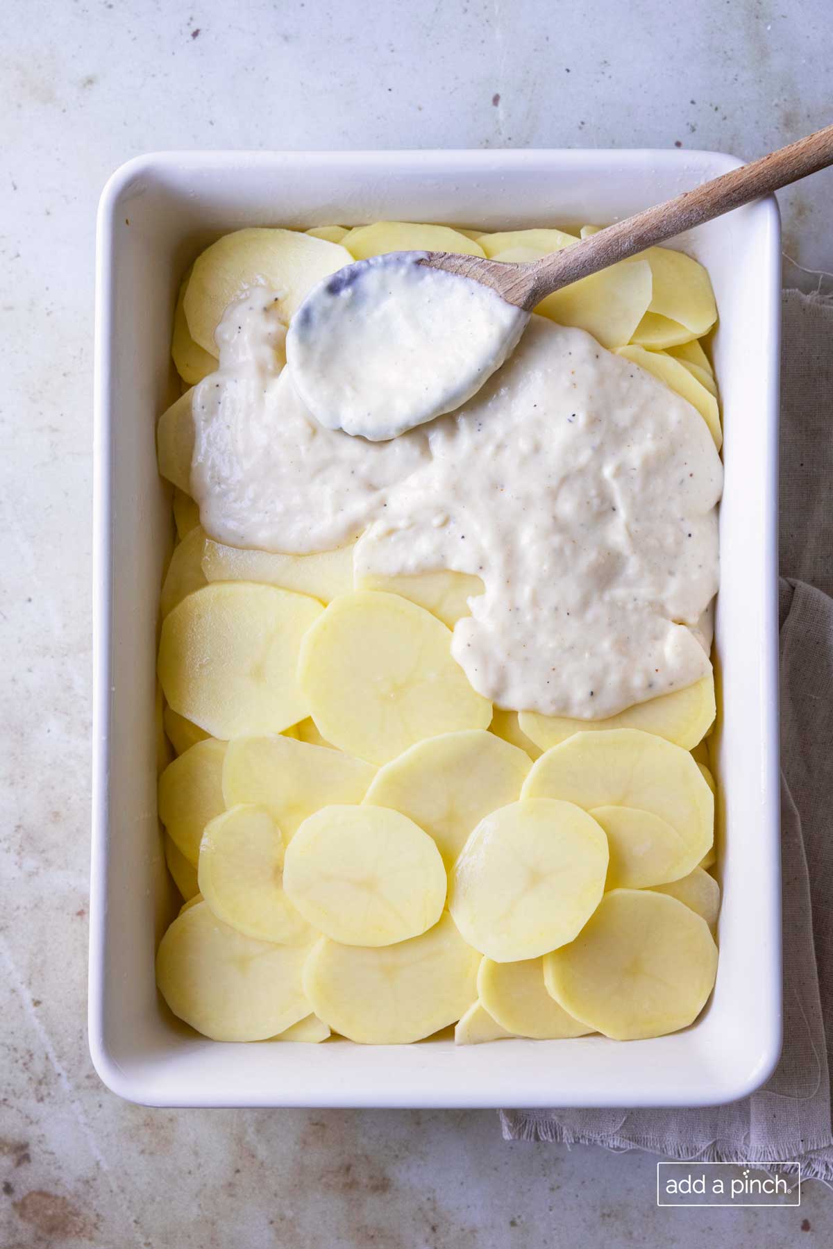 Sliced potatoes in a white baking dish with cream sauce being spread over the top with a wooden spoon.