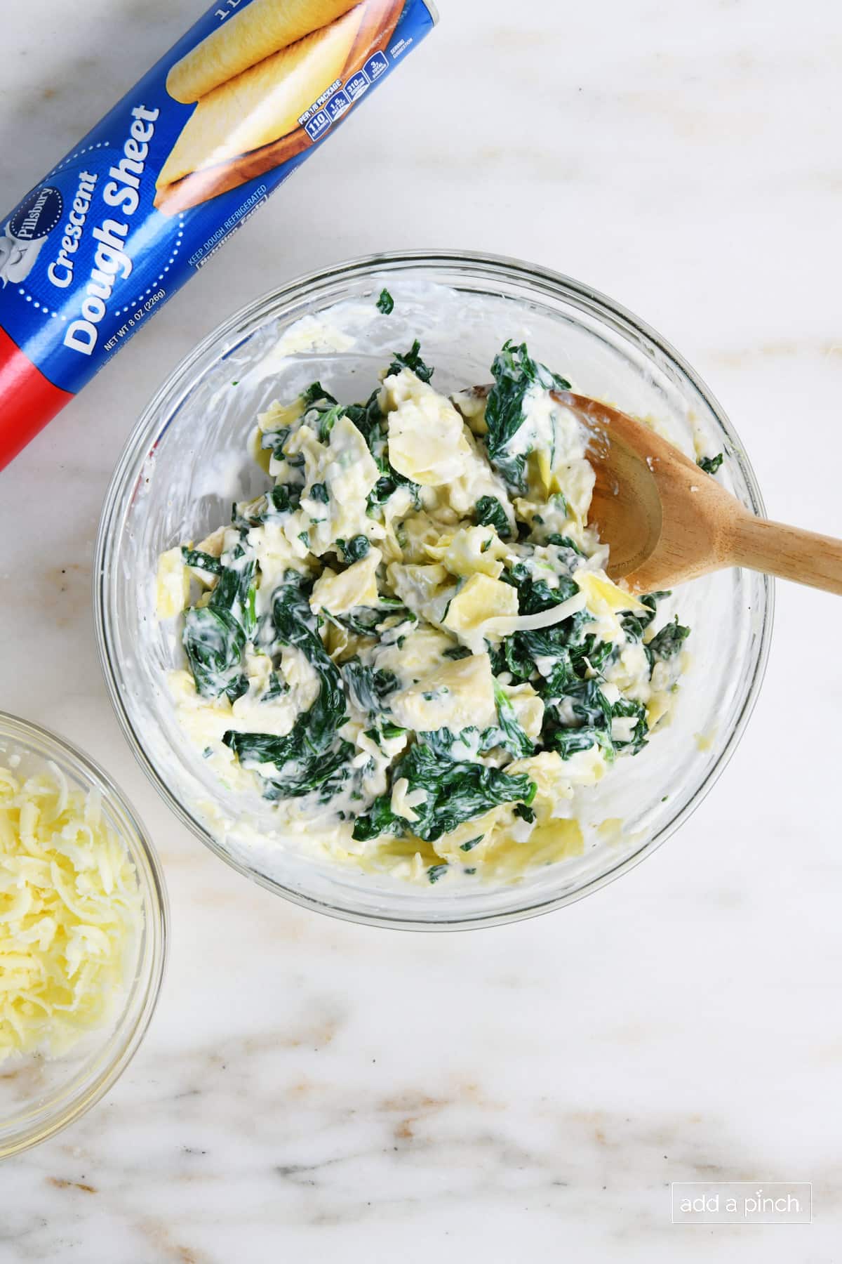 Ingredients for spinach dip bites are mixed in a large mixing bowl with a wooden spoon. A package of crescent dough and a bowl of cheeses sits nearby.