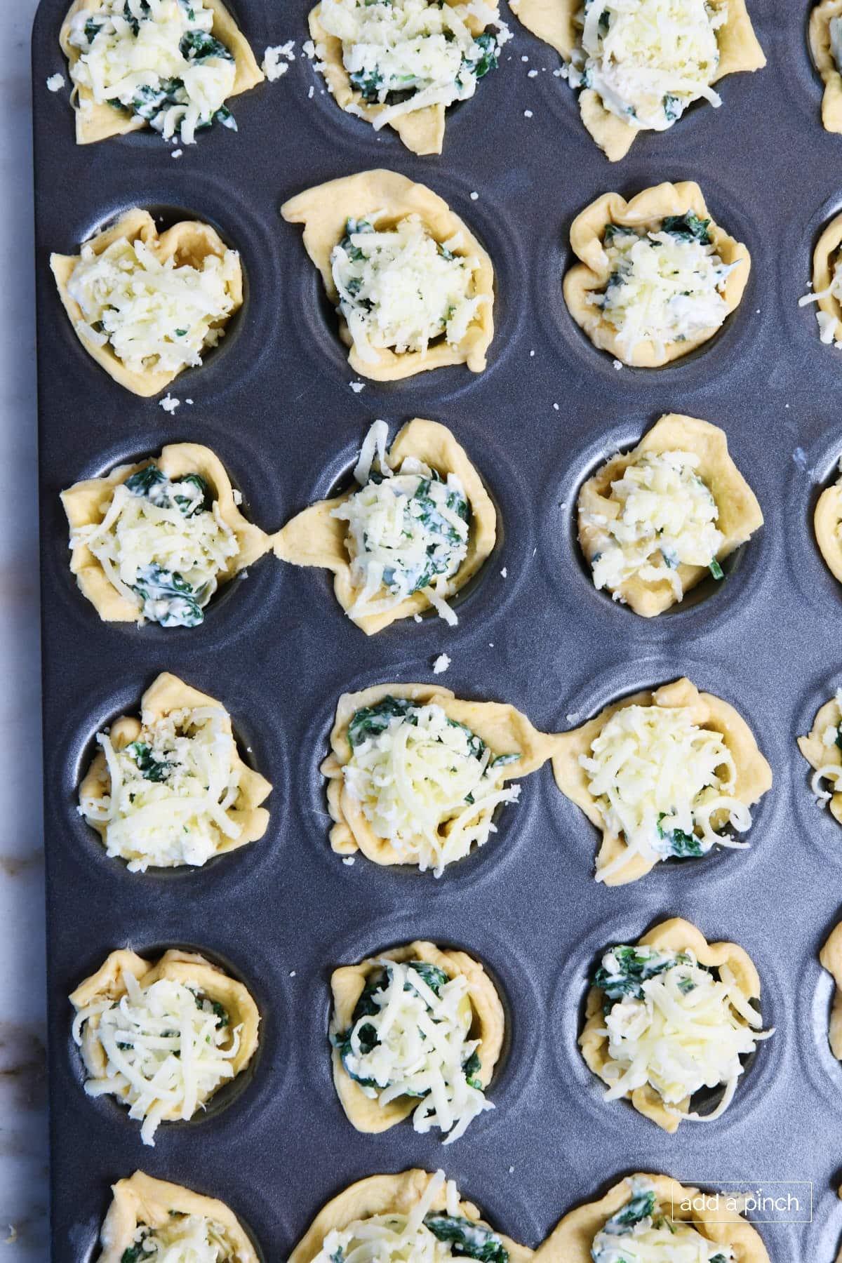 Mini muffin tin holds spinach dip bites freshly topped with cheeses and ready to bake.