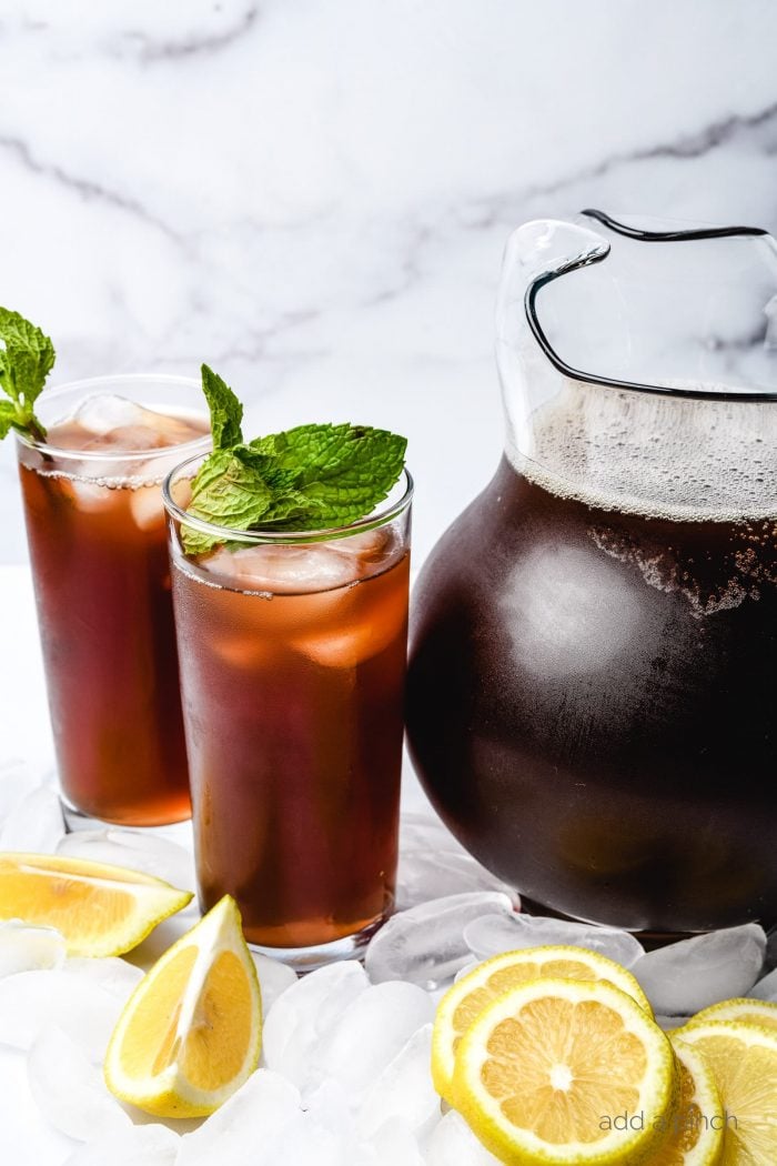 Pitcher of cold sweet tea with two iced tea glasses with sprigs of mint on top, sitting on ice cubes surrounded by lemon slices. 