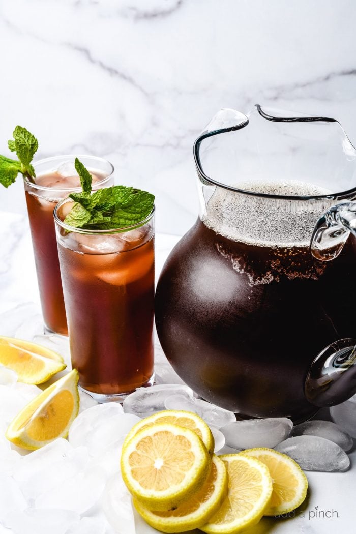 Lemon slices, two glasses of iced tea with mint sprigs beside a glass gallon pitcher of tea. 